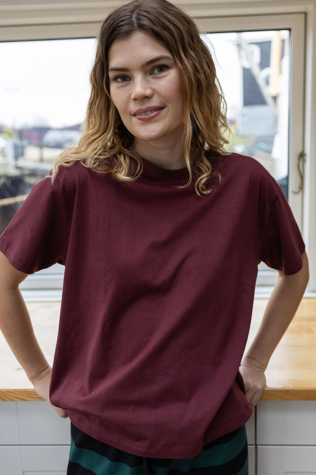 A woman wearing an oversized short-sleeve crewneck t-shirt in maroon, standing in front of a window.