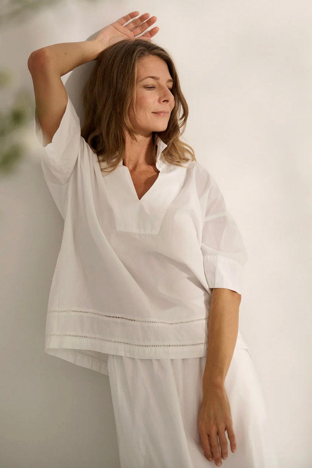 A woman wearing a white, short-sleeved, A-shaped shirt with a detailed hem, posing with one hand behind her head.
