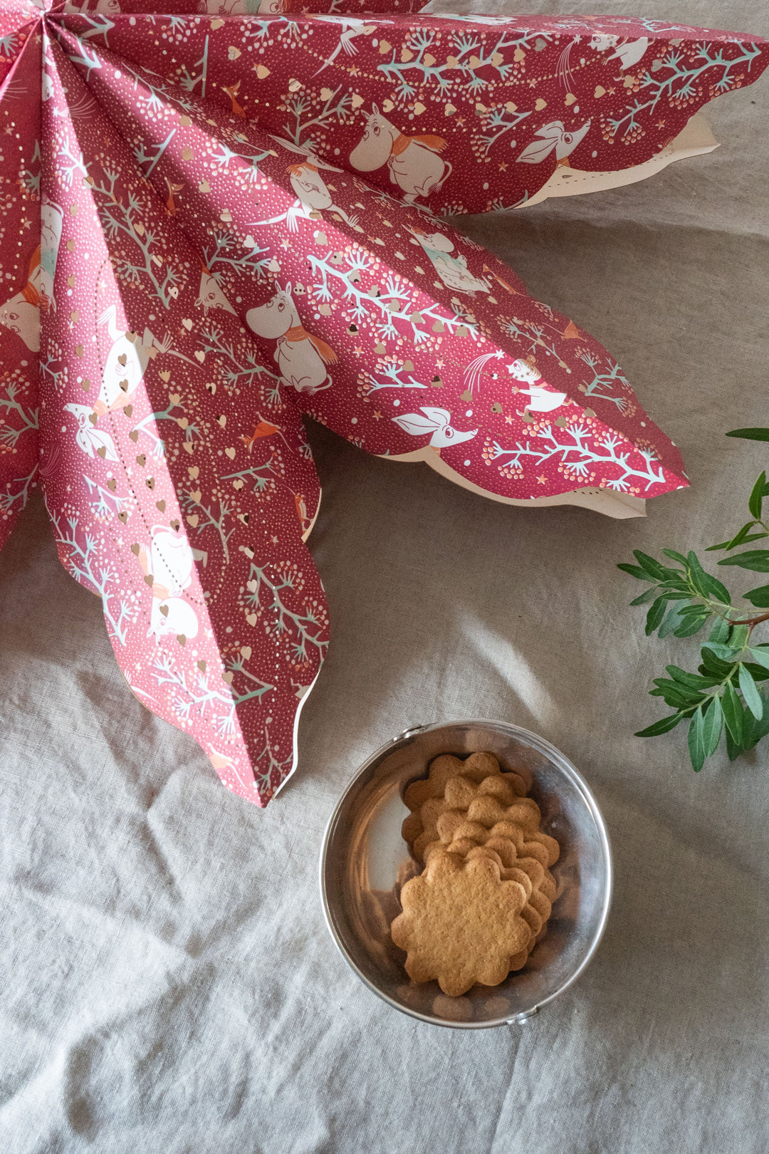 A red paper star with Moomin characters decoration, displayed above a bowl of cookies, with a floral pattern in the background.