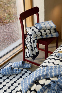 A coordinated bedding and textile set with a bold blue, white, and black scallop pattern, shown folded on a red chair beside an open window, with matching slippers on the floor and a bed in the foreground.