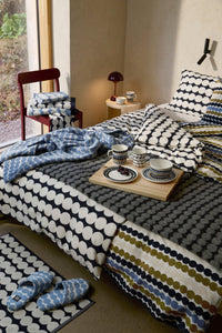 A styled bedroom scene showing a bed with patterned bedding, matching pillows, a folded blue patterned bathrobe, a tray with teacups, a chair with folded towels, and a matching bathmat near slippers. The room has warm neutral walls and natural light from a window.