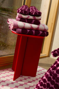 A stack of folded pink and purple patterned towels and a matching pair of slippers displayed on a bright red pedestal in front of a window.