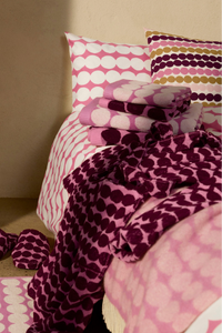 A styled bed with pink and white polka dot bedding, matching folded textiles, and a dark magenta patterned throw blanket, alongside matching slippers on a beige floor against a beige wall.