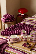 A styled bedroom scene showing coordinated bedding and tableware with a repeating oval dot pattern in purple, pink, olive green, and cream. The bedding is spread across a bed, with matching folded textiles on a small stool, cups and saucers on a wooden tray, and a patterned storage box.