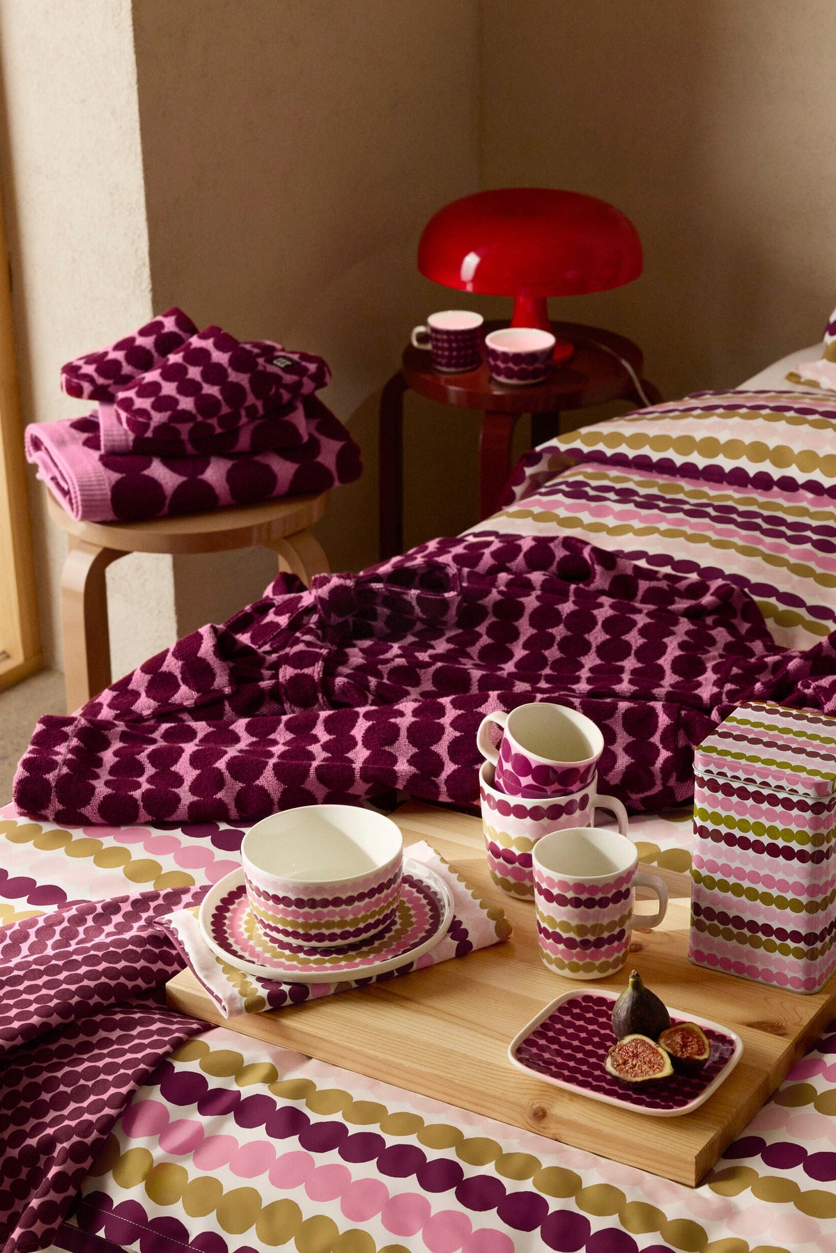 A styled bedroom scene showing coordinated bedding and tableware with a repeating oval dot pattern in purple, pink, olive green, and cream. The bedding is spread across a bed, with matching folded textiles on a small stool, cups and saucers on a wooden tray, and a patterned storage box.