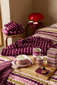 A styled bedroom scene showing coordinated bedding and tableware with a repeating oval dot pattern in purple, pink, olive green, and cream. The bedding is spread across a bed, with matching folded textiles on a small stool, cups and saucers on a wooden tray, and a patterned storage box.