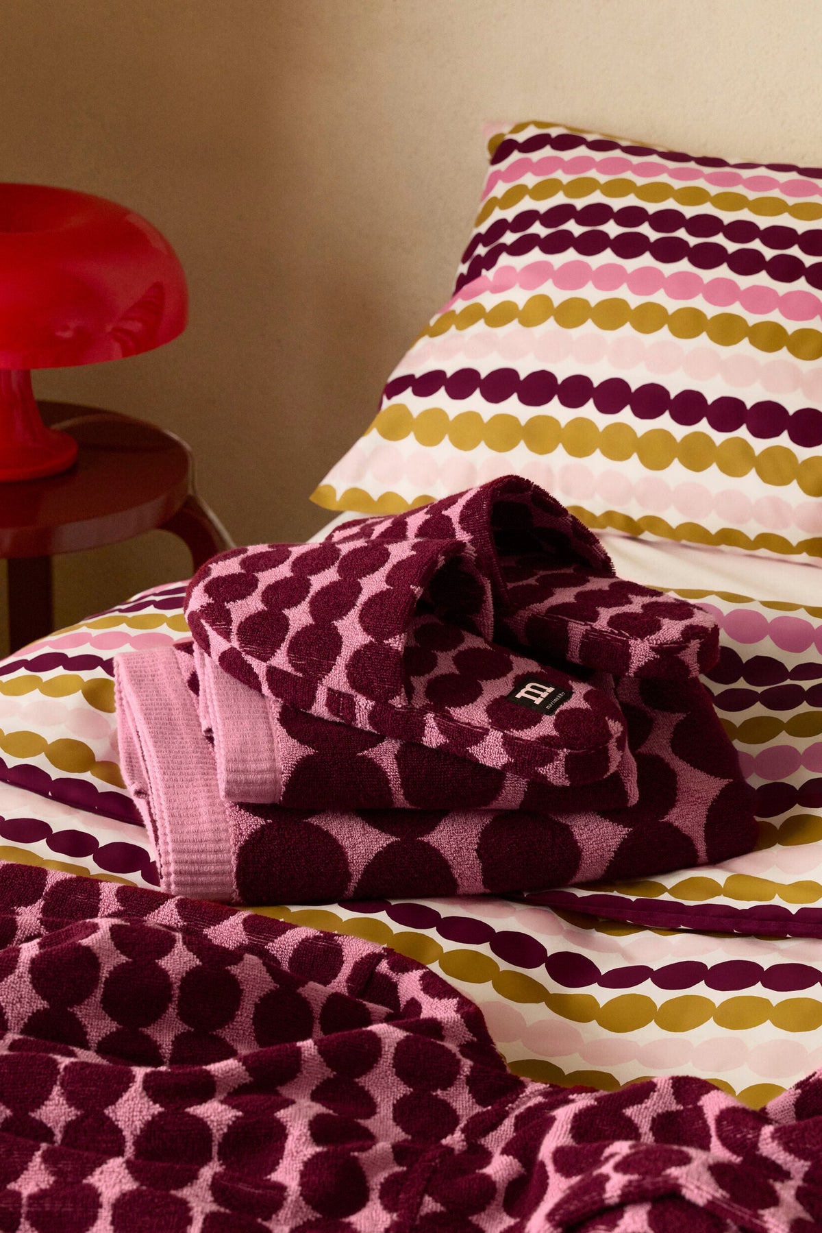 A folded knit blanket and matching pillowcase with horizontal stripes of pink, mustard yellow, and deep burgundy dots on a cream background, styled on a bed next to a red table lamp.