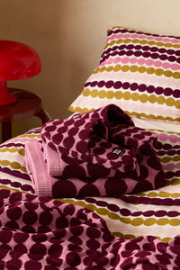 A folded knit blanket and matching pillowcase with horizontal stripes of pink, mustard yellow, and deep burgundy dots on a cream background, styled on a bed next to a red table lamp.