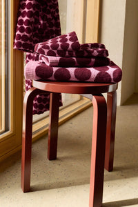 A folded stack of plush bath towels and a matching hanging towel with a dark plum and light pink polka dot pattern, styled on a reddish-brown wooden stool in a sunlit room.