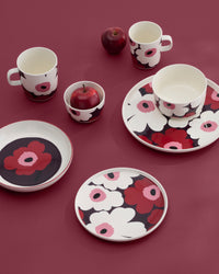 A coordinated floral tableware set arranged on a solid burgundy background, including plates, bowls, and cups with a bold flower pattern in white, pink, and red. Two red apples are placed near the set as styling props.