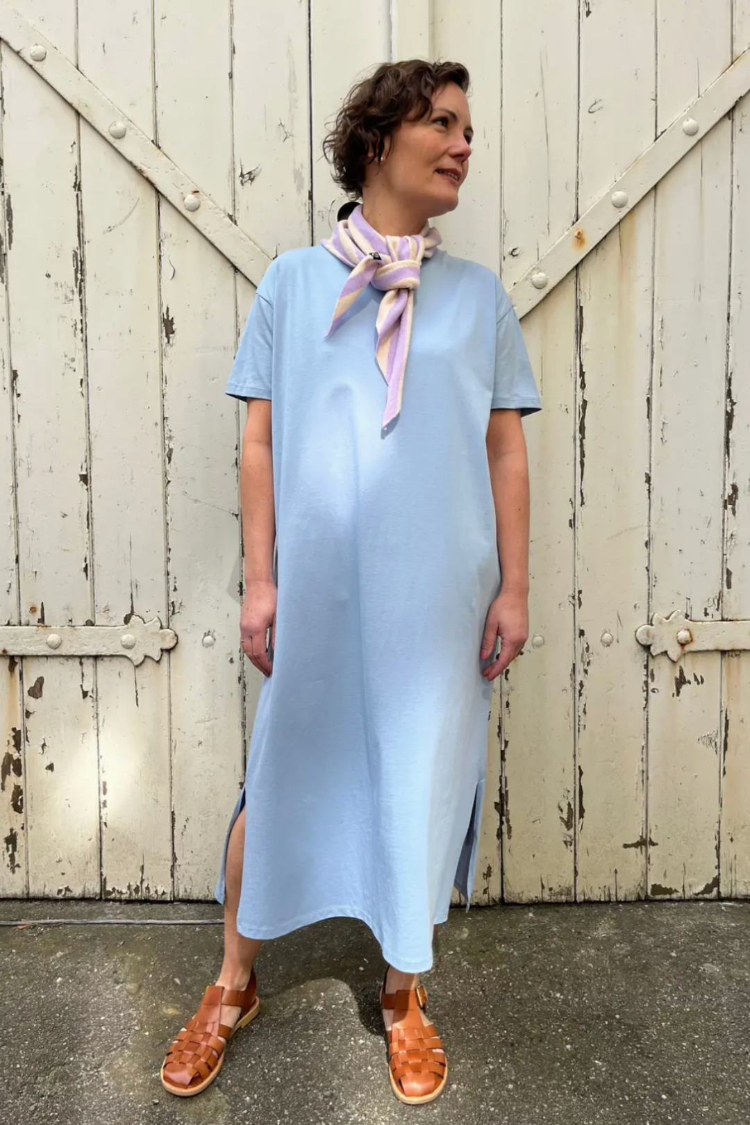 Woman wearing a light blue dress with a pink scarf, standing in front of a rustic wooden door.