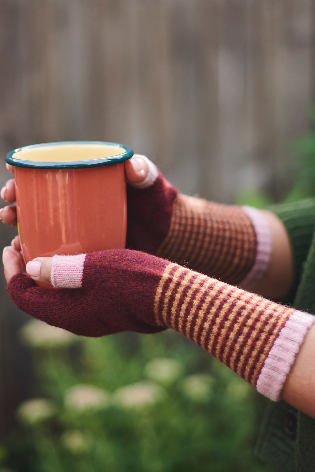 Person wearing striped gloves holding a terracotta-colored cup outdoors.